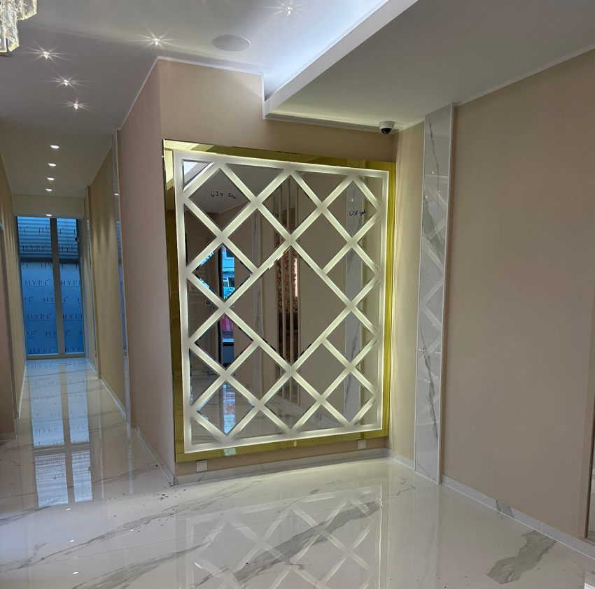 Illuminated geometric lattice mirror with a polished gold frame installed in a modern hallway with marble flooring.
